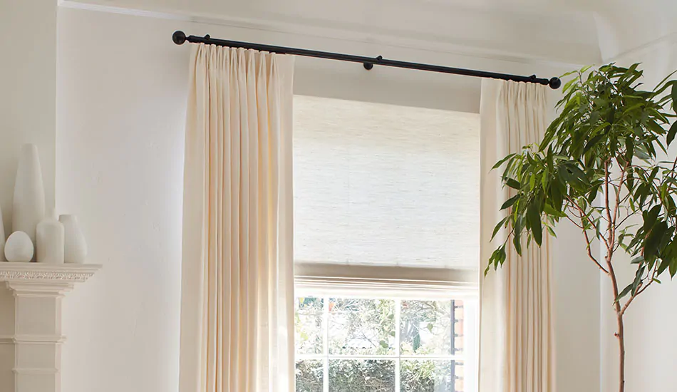 A living room window has Tailored Pleat Drapery made of Luxe Linen in Oyster and a woven shade made of Harper in Ivory