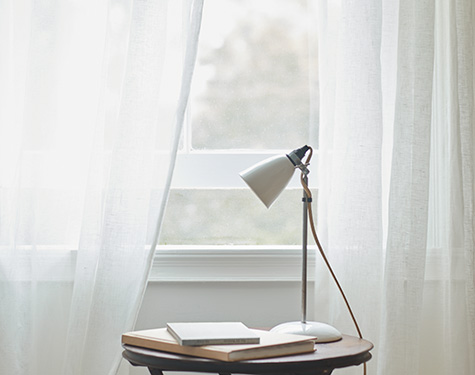 A small round end table sits near a window with one of the types of drapes material, sheer drapes in an off-white color