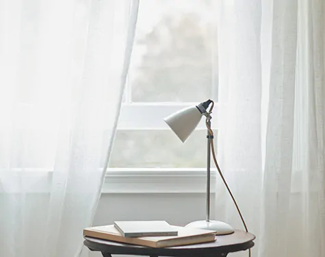 A small round end table sits near a window with one of the types of drapes material, sheer drapes in an off-white color