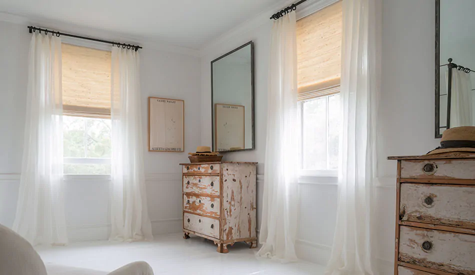 A bedroom with rustic furniture features curtain shades made of Luxe Sheer Linen in Off-White layered with Woven Wood Shades