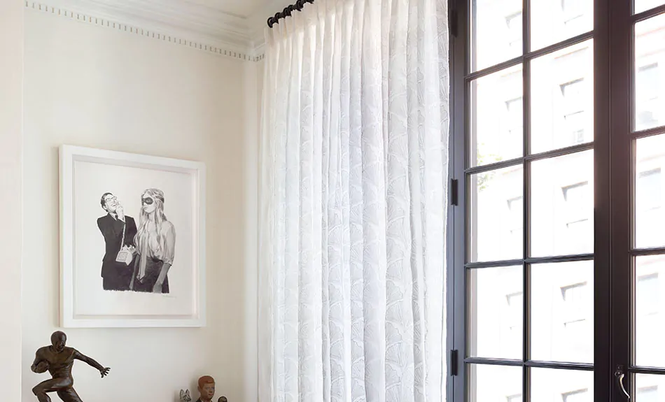 A modern sitting room features tall Tailored Pleat Drapery made of The Novogratz Feather Palm Embroidery in Vintage Lace