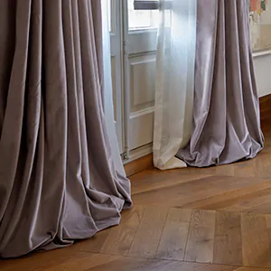 Tailored Pleat Drapery made of Posh Velvet in Heather puddles on the warm wood floor with a herringbone pattern of a bedroom