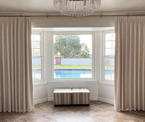 A bay window in a room with a luxe chandelier has Velvet curtains in the Tailored Pleat style made of Velvet in White