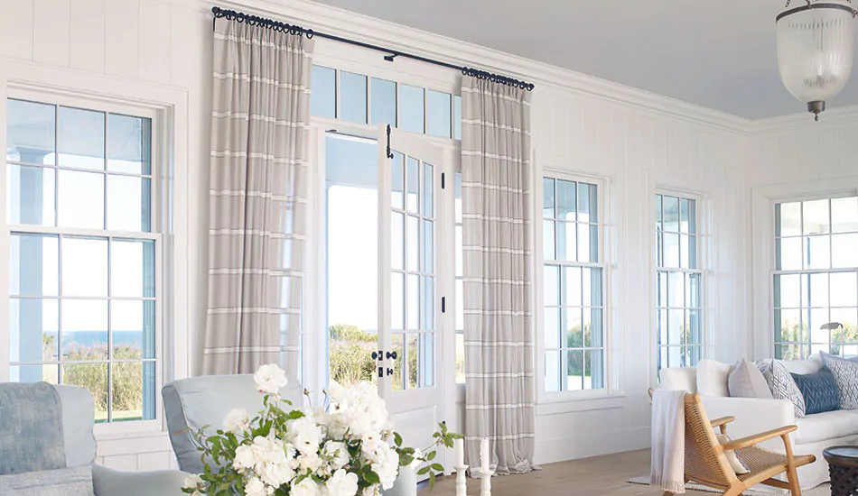 A bright living room has white walls and light blue accents along with Tailored Pleat Drapery made of Harbor Stipe in Sand