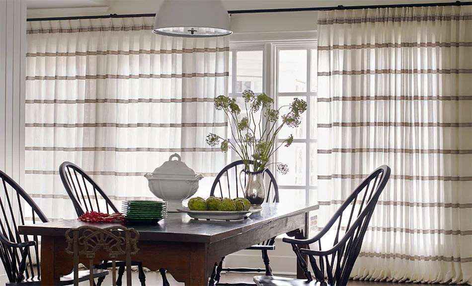 A rustic dining room features large windows covered by curtain sheers made of Victoria Hagan's Harbor Stripe in Stone
