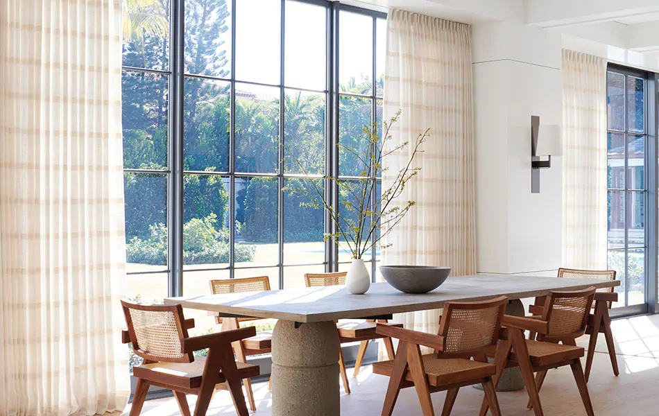 A dining room has window treatments for large windows made of Tailored Pleat Drapery in Lily, Buff which adds warmth