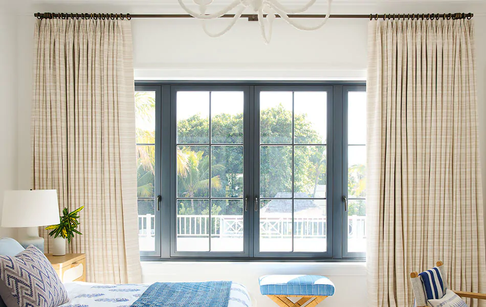 Beige-sand-colored curtains are pulled apart to reveal trees and a pool while inside light falls on a crisply made bed