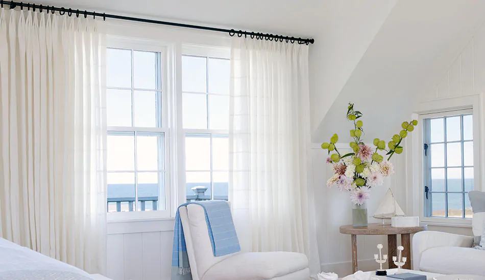 A bright coastal bedroom features Tailored Pleat Drapery made from Sankaty Stripe in Moon for a light airy look