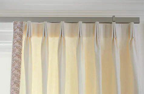 A close up of cream colored tailored pleat drapery shows curtains hung on a track system for easy functionality