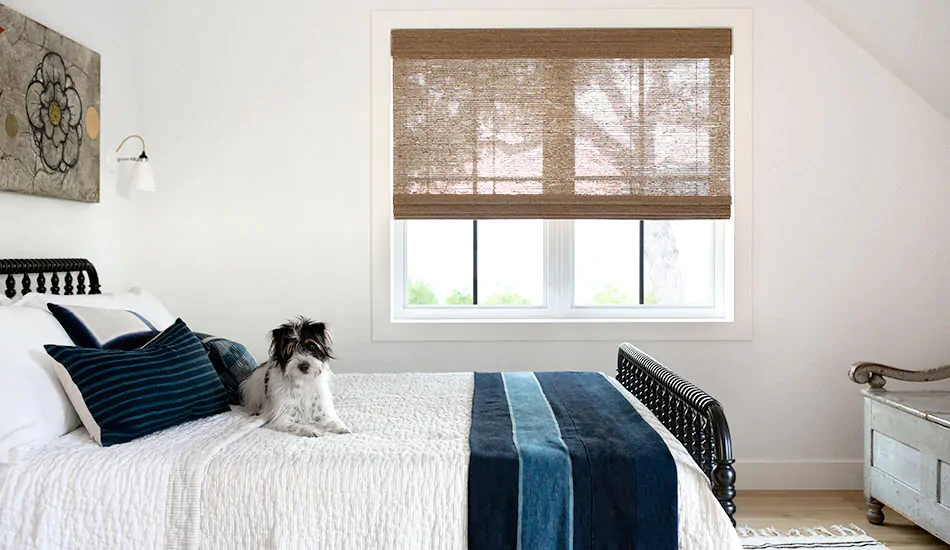 A dog sits on a bed in a bedroom with a Waterfall Woven Wood Shade made of Artisan Weaves Bayshore, Sage for an organic look