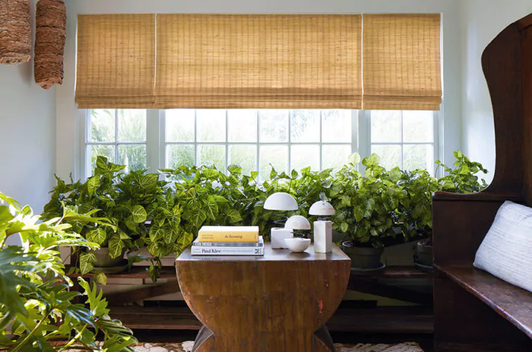 Sunroom window treatments, woven wood shades made of Coastline in Oat cover windows in a sunroom filled with lush greenery