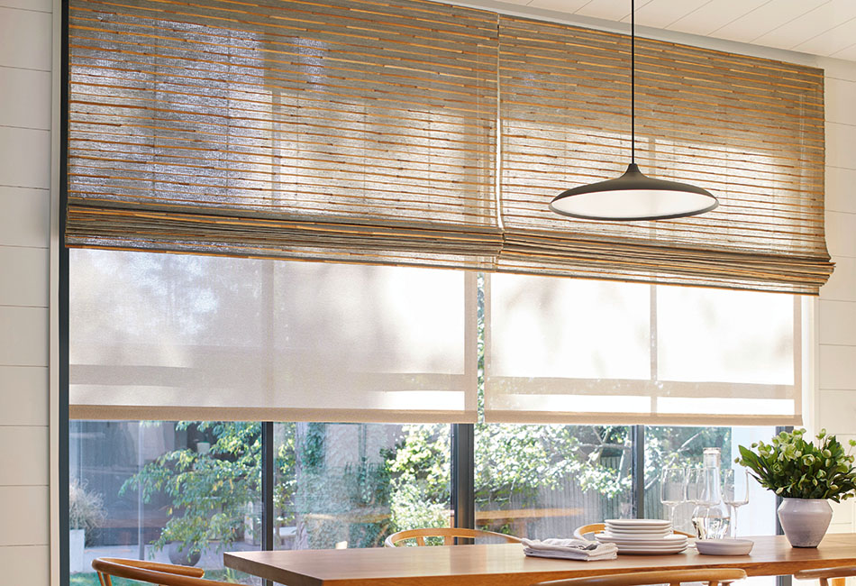 A dining room with a wood table has woven shades made of Artisan Weaves Reed in Slate and Roller Shades of Sullivan in Sand