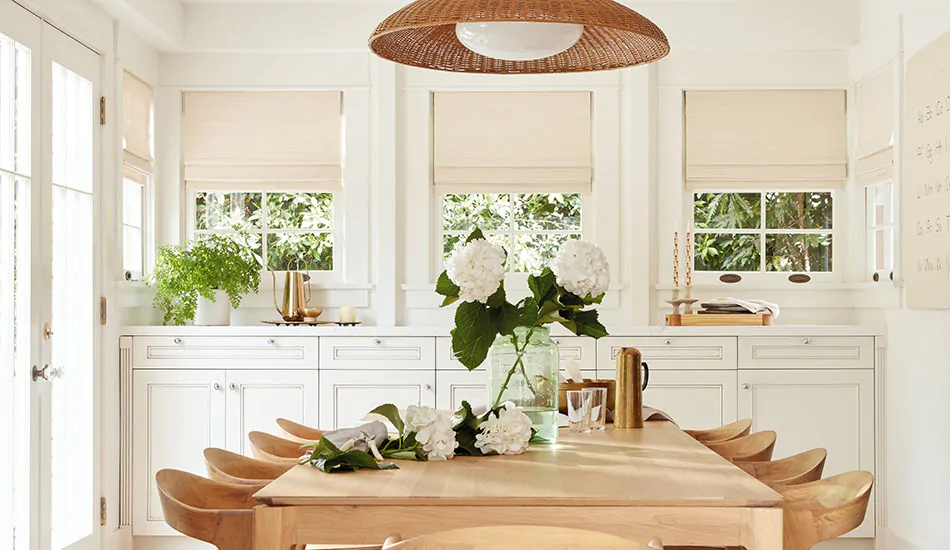 A bright boho-inspired kitchen features Woven Wood Shades made of Somerset in Cloud and a long light wood dining table