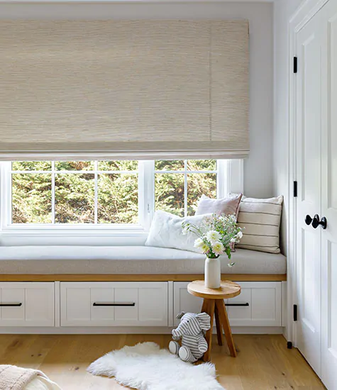 A room with a plush window seat has blackout shades for nursery windows made of Waterfall Woven Wood Shade, Grassweave, Hemp