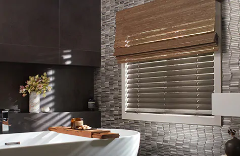 A bathroom has layered window treatments of Woven Wood Shades of Del Rey in Taupe and Wood Blinds of Bamboo in Black
