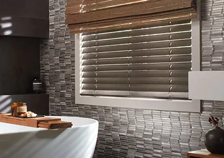 A bathroom with dark tiling, a white tub and wood accents has a window with 2-inch Bamboo blinds in Black