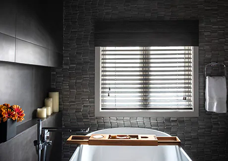 A bathroom with dark tiling, a white tub and wood accents has a window with 2-inch Bamboo blinds in Black