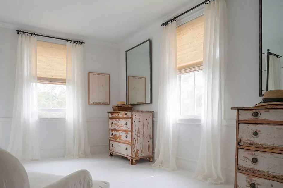 A main bedroom features sheer curtains and woven wood shades which are one of the types of shades for windows
